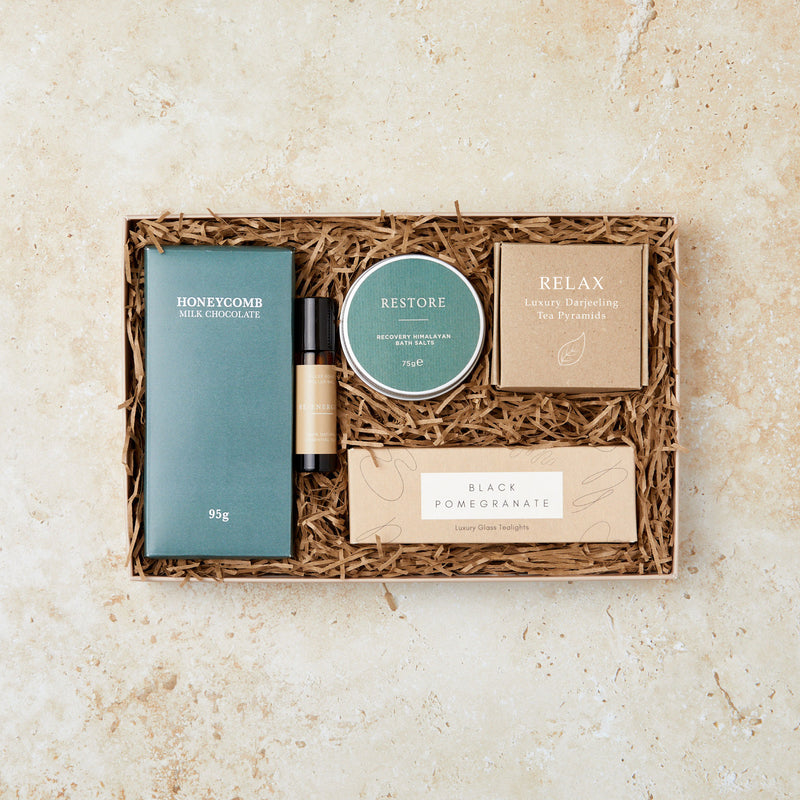 Relaxation gift set containing honeycomb chocolate bar, restore bath salts, darjeeling tea, aromatherapy oil roll on and black pomegranate tealights in green and kraft packaging