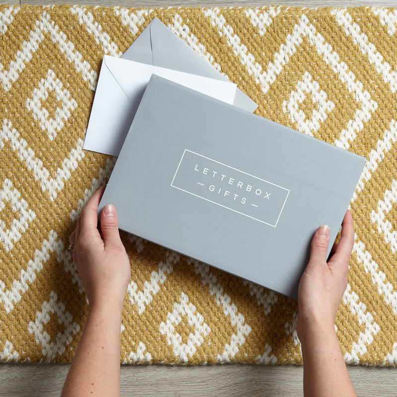 someone picking up a letterbox friendly gift box from their door mat. a grey gift box with the Letterbox Gifts logo