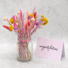 Bright summer dried flower bouquet in pinks, purples and yellows arranged in a glass jar next to a congratulations greetings card