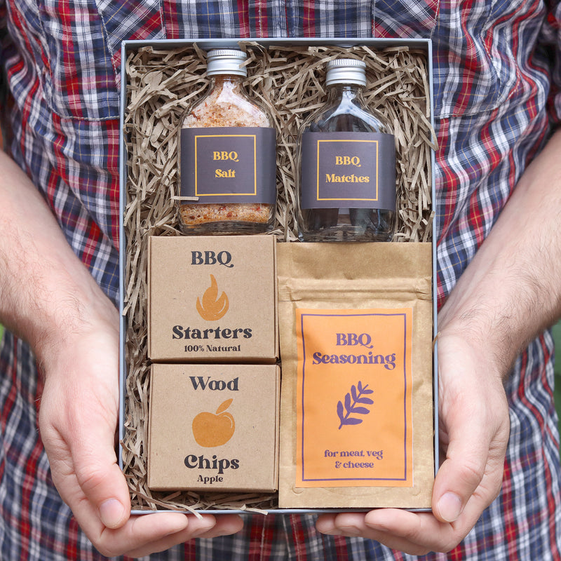 A man in checkered shirt holding a BBQ gift set containing BBQ salt, matches, BB! starters, wood chips and BBQ seasoning in Kraft design