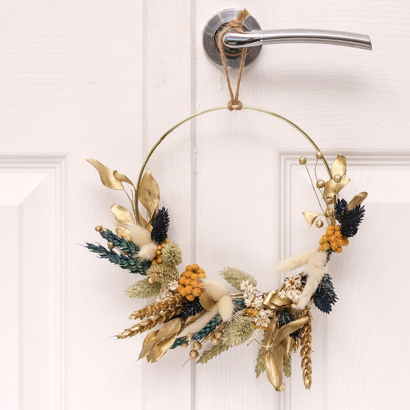 Dried flower wreath constructed using blue, yellow, gold & natural dried flowers on a gold hoop, hanging on a white door