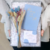 A person holding an engagement gift set containing a blue wedding-planner notebook, tea for two Darjeeling tea box and dried flower posy