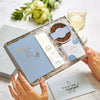 A person holding an Engagement gift set in blue and white containing wedding planner notebook, chocolate honeycomb, Darjeeling tea for two box and You're Engaged tealights