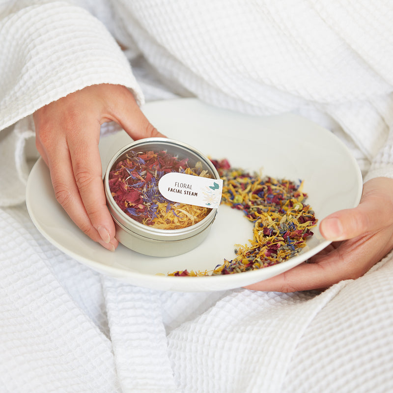 A person in a bath robe holding a floral facial steam tin containing red, blue and yellow flower petals