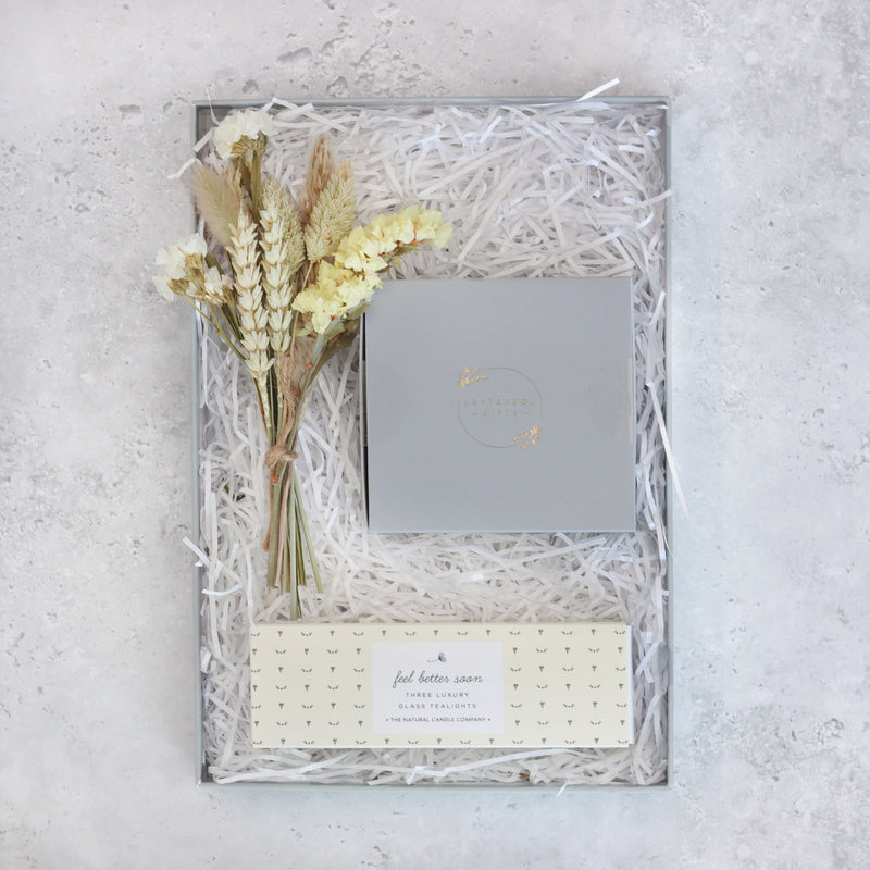 top view of a gift box containing a neutral coloured bouquet of flowers, a box of butterscotch chocolates and box of candles with 'feel better soon' packaging