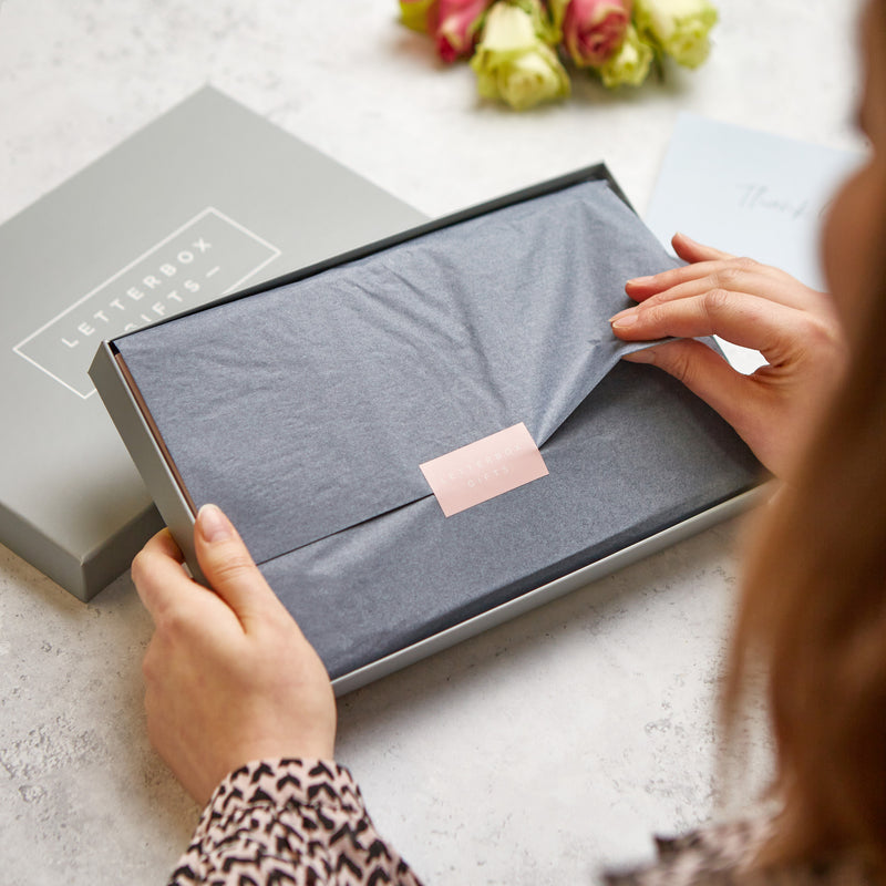 a woman sat at a table opening a grey letterbox gift box, peeling the grey gift wrapping and letterbox gift logo on the sticker