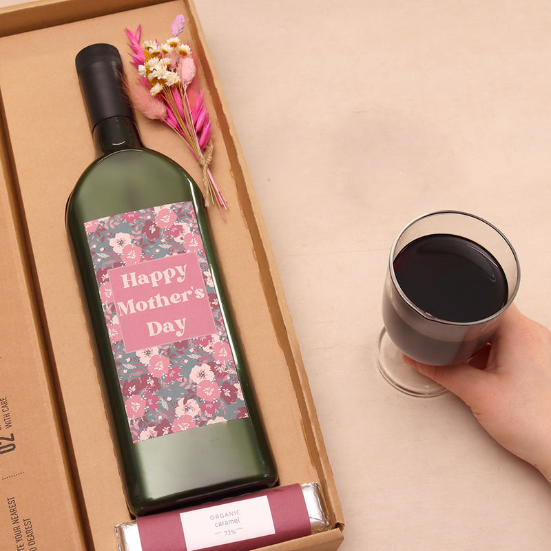 A person enjoying a glass of red wine alongside their letterbox-friendly mother's day wine bottle, sweet orange caramel  choc & dried flower posy
