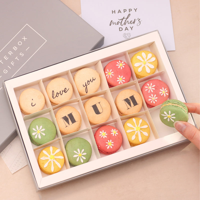 Fifteen mother's day macarons in pastel pink, green, yellow and cream, with 'i love you mum' printed text and white daisies