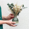 A person in green shirt holding a neutral-tone dried flower bouquet arranged in a green ombre vase