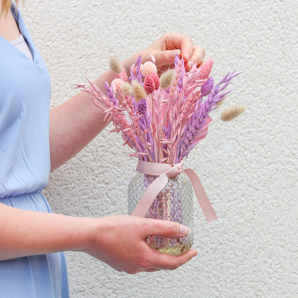 A person in blue dress holding a pastel pink and purple dried flower bouquet arranged in a glass jar