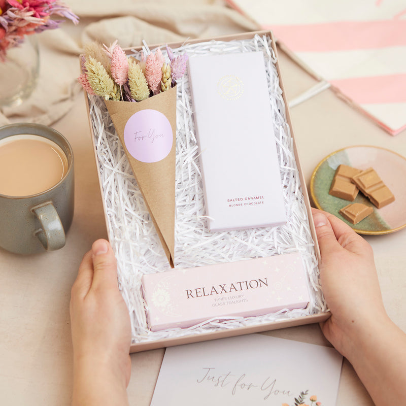 A person holding their Relaxation letterbox gift set containing purple relaxation tealights, salted caramel chocolate bar & pastel dried flower posy