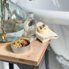 Bath essence bottle, cocoa butter massage bar and floral facial steam tin next to a bathtub