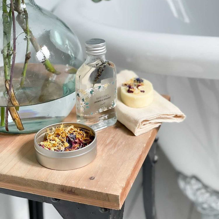 Bath essence bottle, cocoa butter massage bar and floral facial steam tin next to a bathtub
