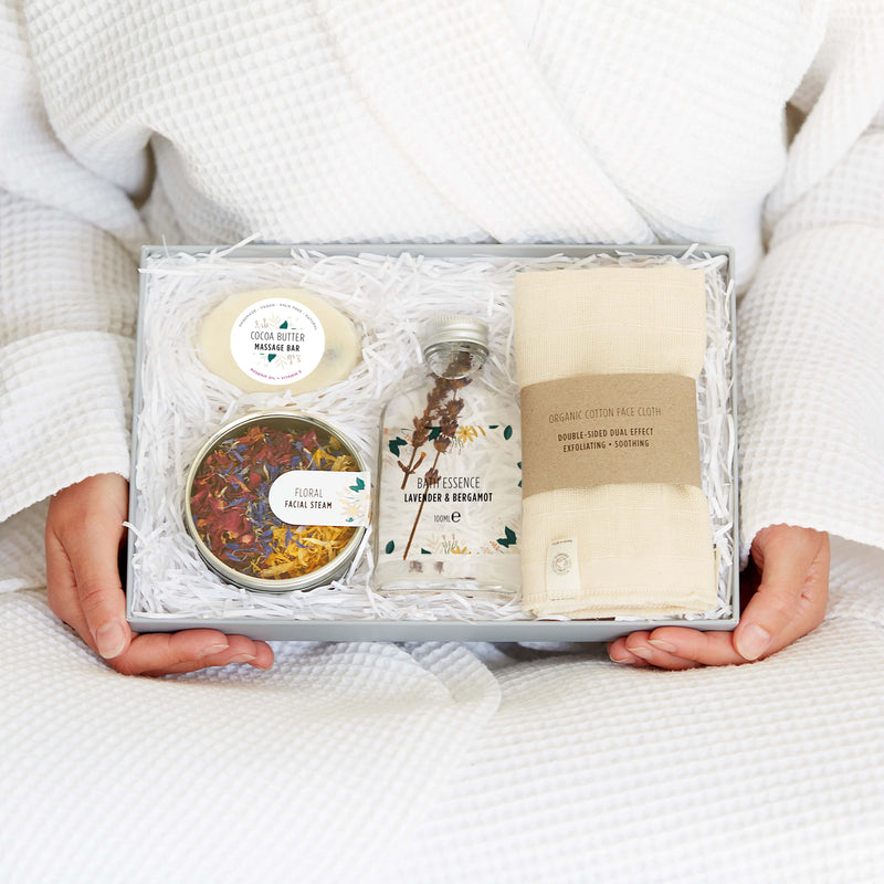 A person in spa robe holding an eco friendly spa gift set containing organic cotton face cloth, bath essence, floral facial steam tin and cocoa butter massage bar