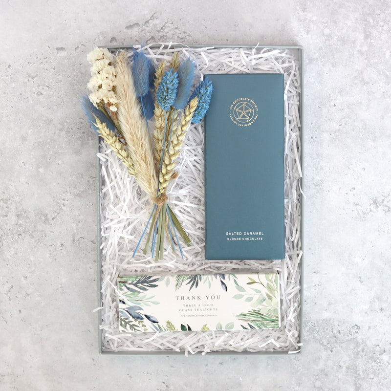 top view of a gift box containing a bar of chocolate in dark blue card packaging, a box of tealights with a green  plant pattern with the words 'thank you', and a bouquet of dried flowers