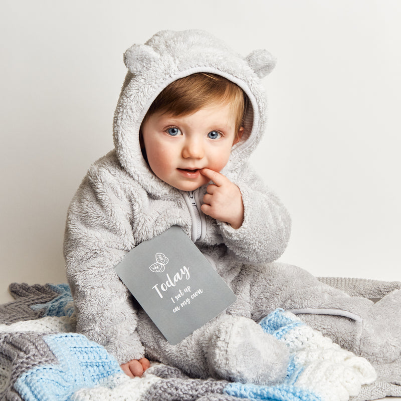 A baby in fluffy grey jumpsuit next to a baby milestone card reading 'Today I sat up on my own'