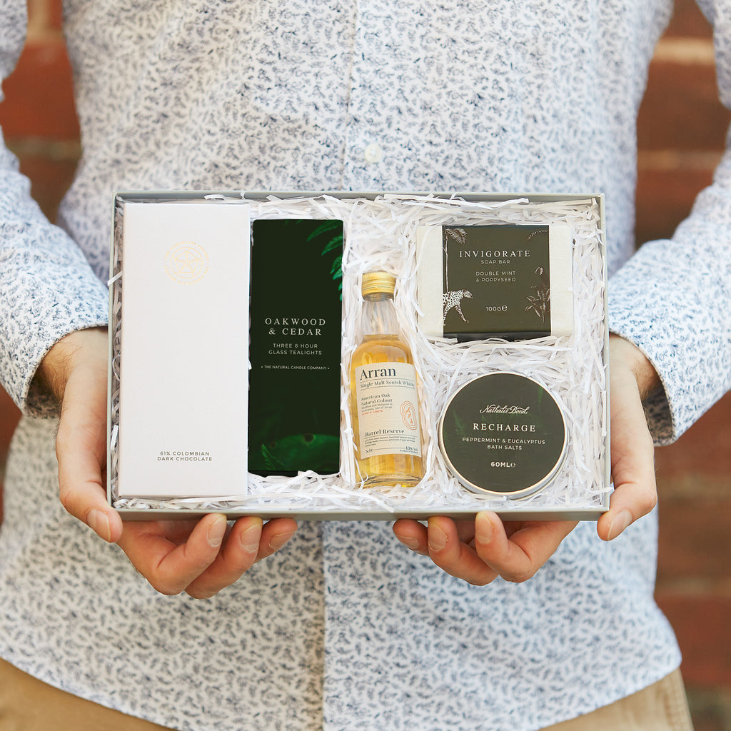 A man holding their men's letterbox gift set containing chocolate bar, oakwood & cedar tealights, whisky, soap bar and bath salts