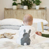 A baby sitting up in bed wearing a white baby grow with grey bunny rabbit image