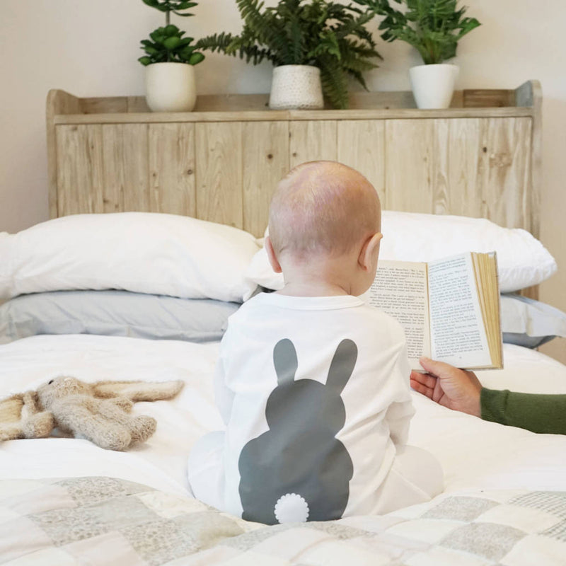 A baby sitting up in bed wearing a white baby grow with grey bunny rabbit image