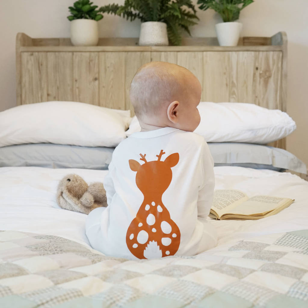 A baby sitting on a bed in a white baby grow with orange fawn image on the back